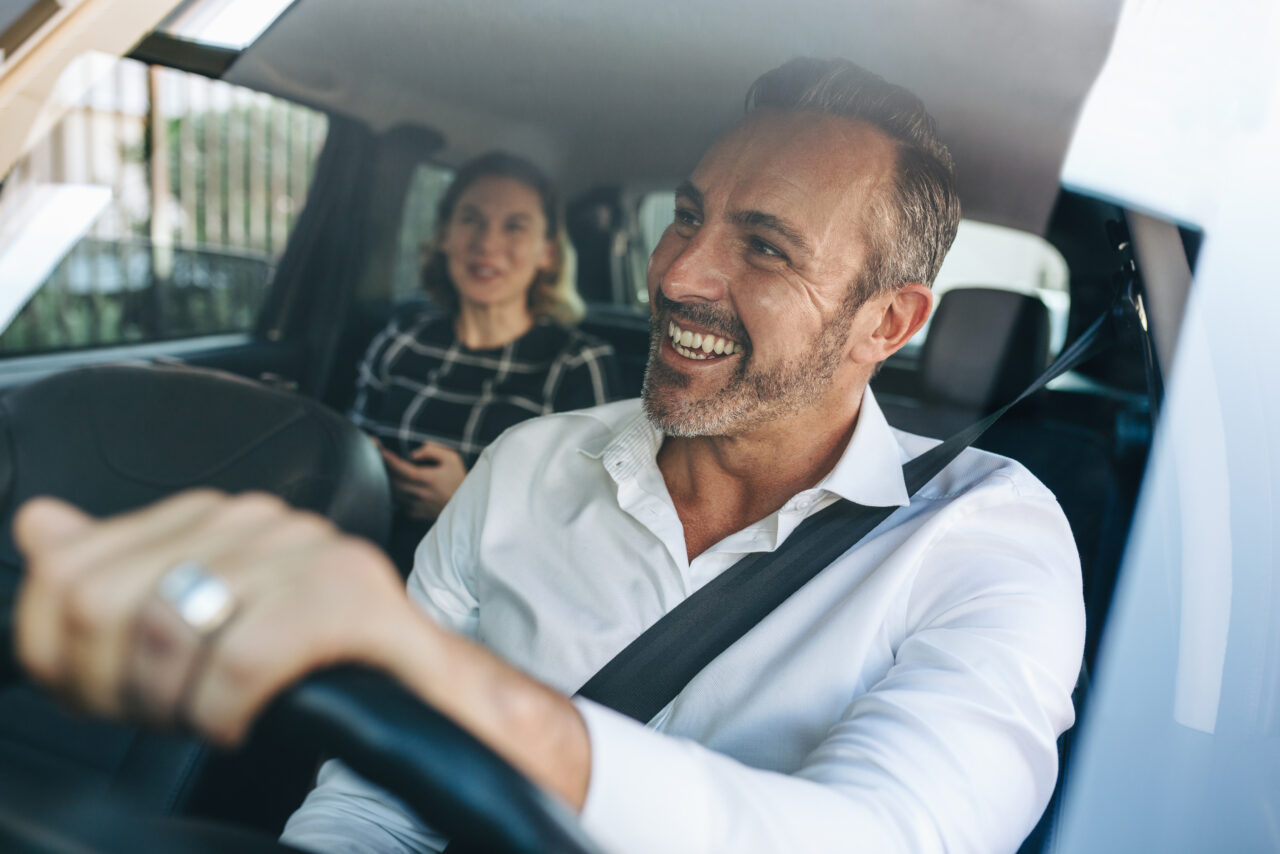 Chauffeur Care 4 U Logistics au volant, souriant, avec une passagère à l'arrière lors d'un transport personnalisé en Belgique